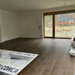 Wnętrze w trakcie remontu z ułożoną, ciemną podłogą z paneli, widoczne materiały budowlane, okno z widokiem na ogr&oacute;d z trampoliną.