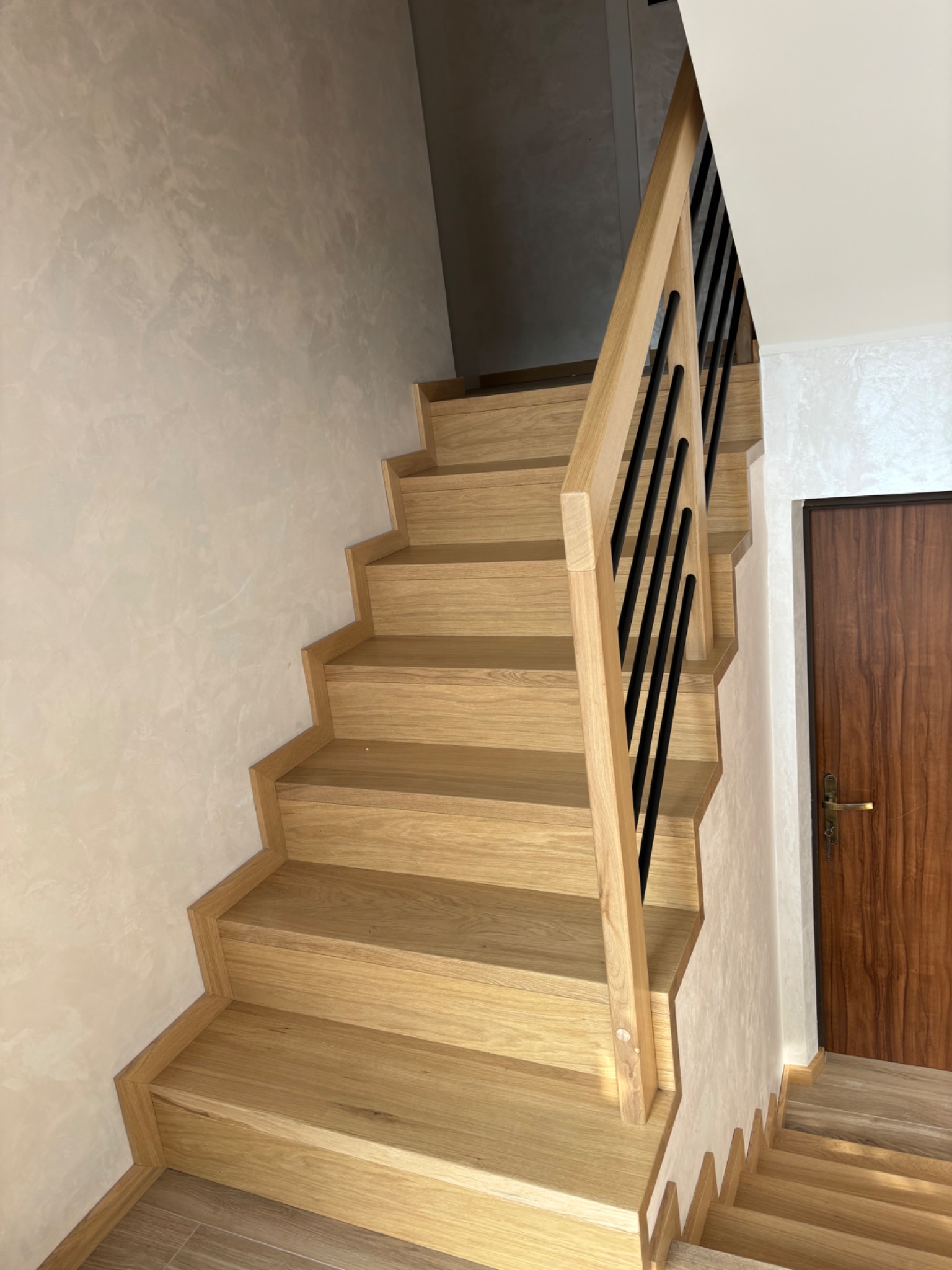 Jasne drewniane schody z czarnymi metalowymi prętami balustrady, widok z dołu, ściana z dekoracyjnym tynkiem.