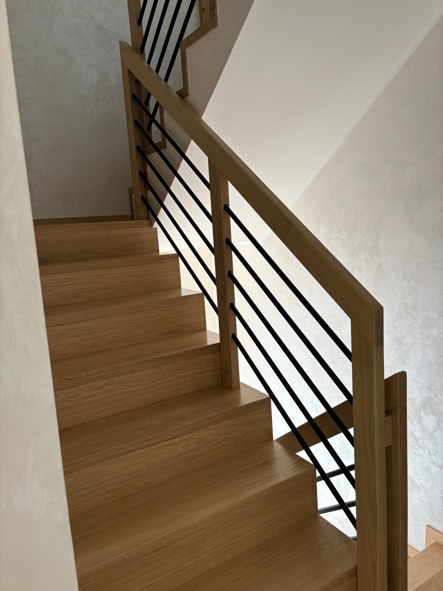 Jasne drewniane schody z czarnymi, metalowymi balustradami w nowoczesnym domu z kremowymi ścianami o fakturze trawertynu.