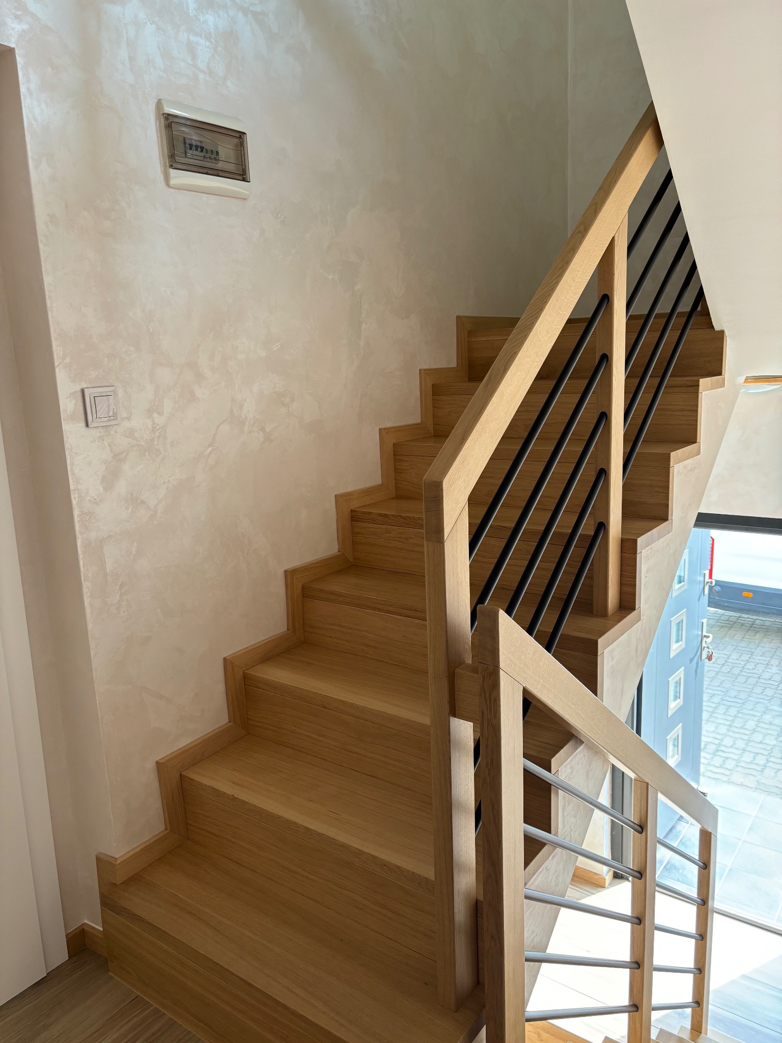 Jasne, drewniane schody z czarnymi metalowymi prętami balustrady, widok z dołu, ściana wykończona w stylu trawertynu, skrzynka elektryczna na ścianie.