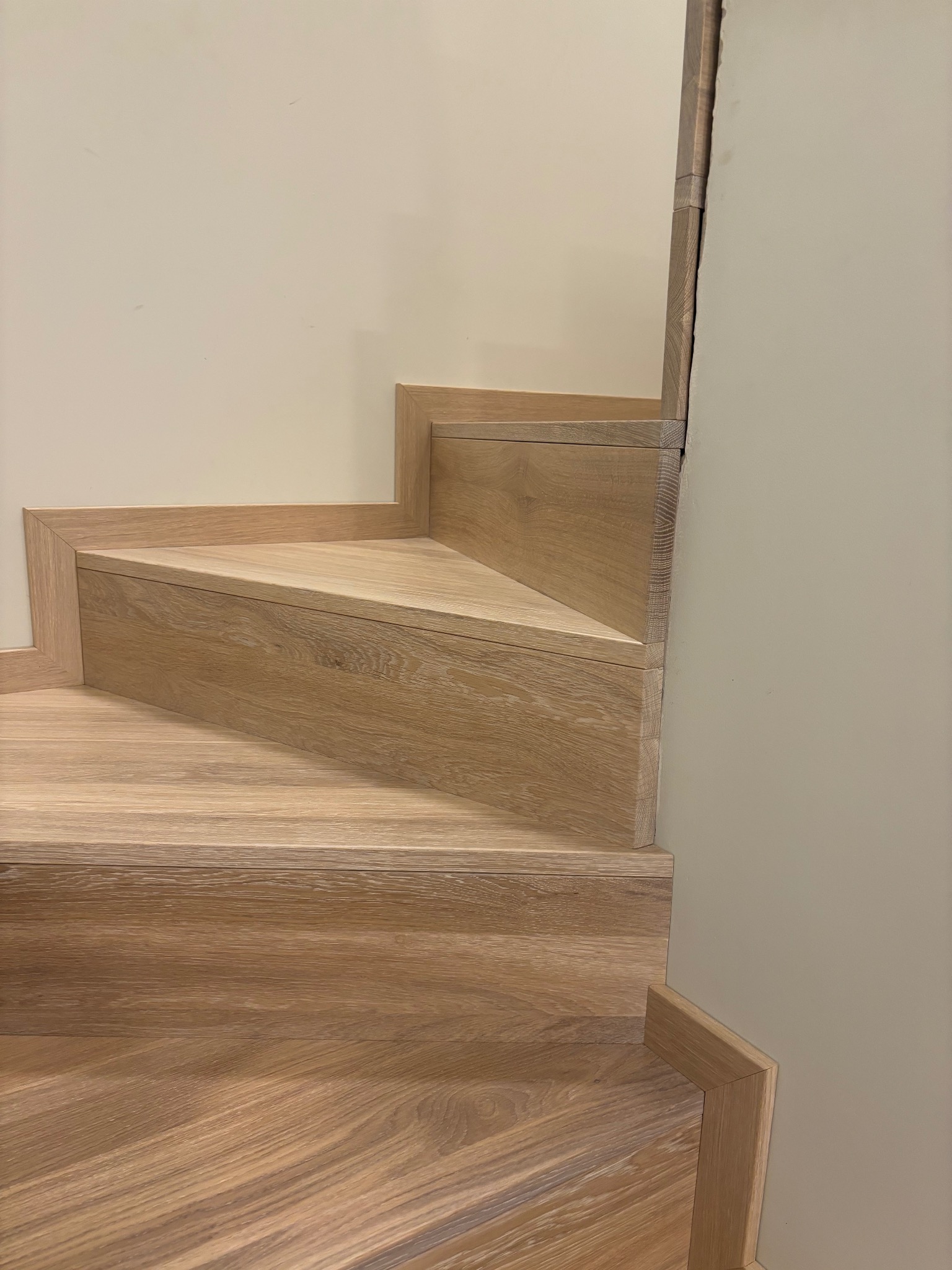 Fragment drewnianych schodów z jasnego drewna, widoczne dwa stopnie z triangularnym podestem, przy ścianie w kolorze złamanej bieli.