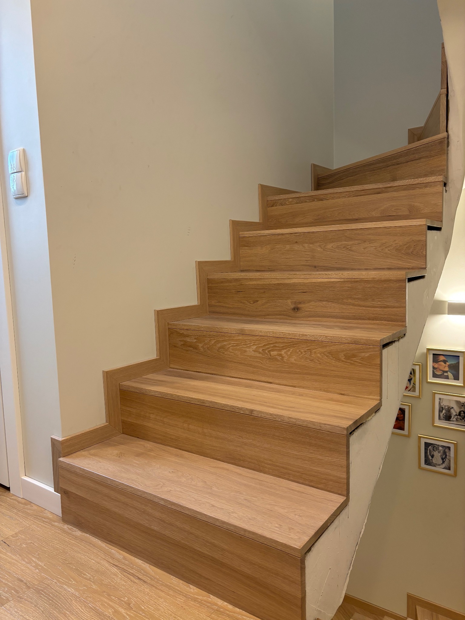 Jasne, dębowe schody z widocznymi słojami drewna, wznoszące się wzdłuż ściany z ramkami ze zdjęciami, oświetlone ciepłym światłem.