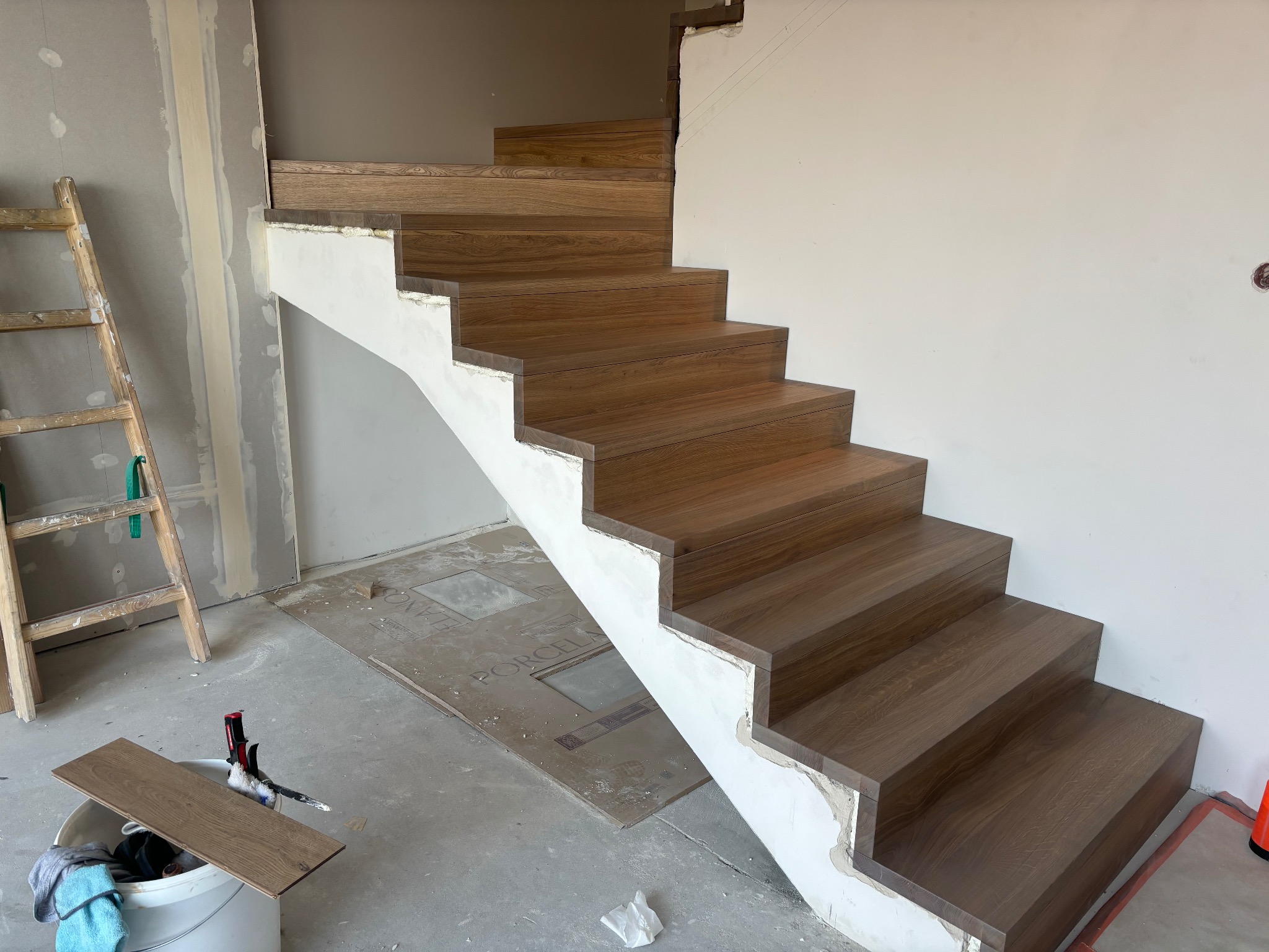 Montaż drewnopodobnych stopni schodowych na betonowej konstrukcji, widoczna drabina i narzędzia w wiadrze, ślady prac remontowych na ścianach i podłodze.