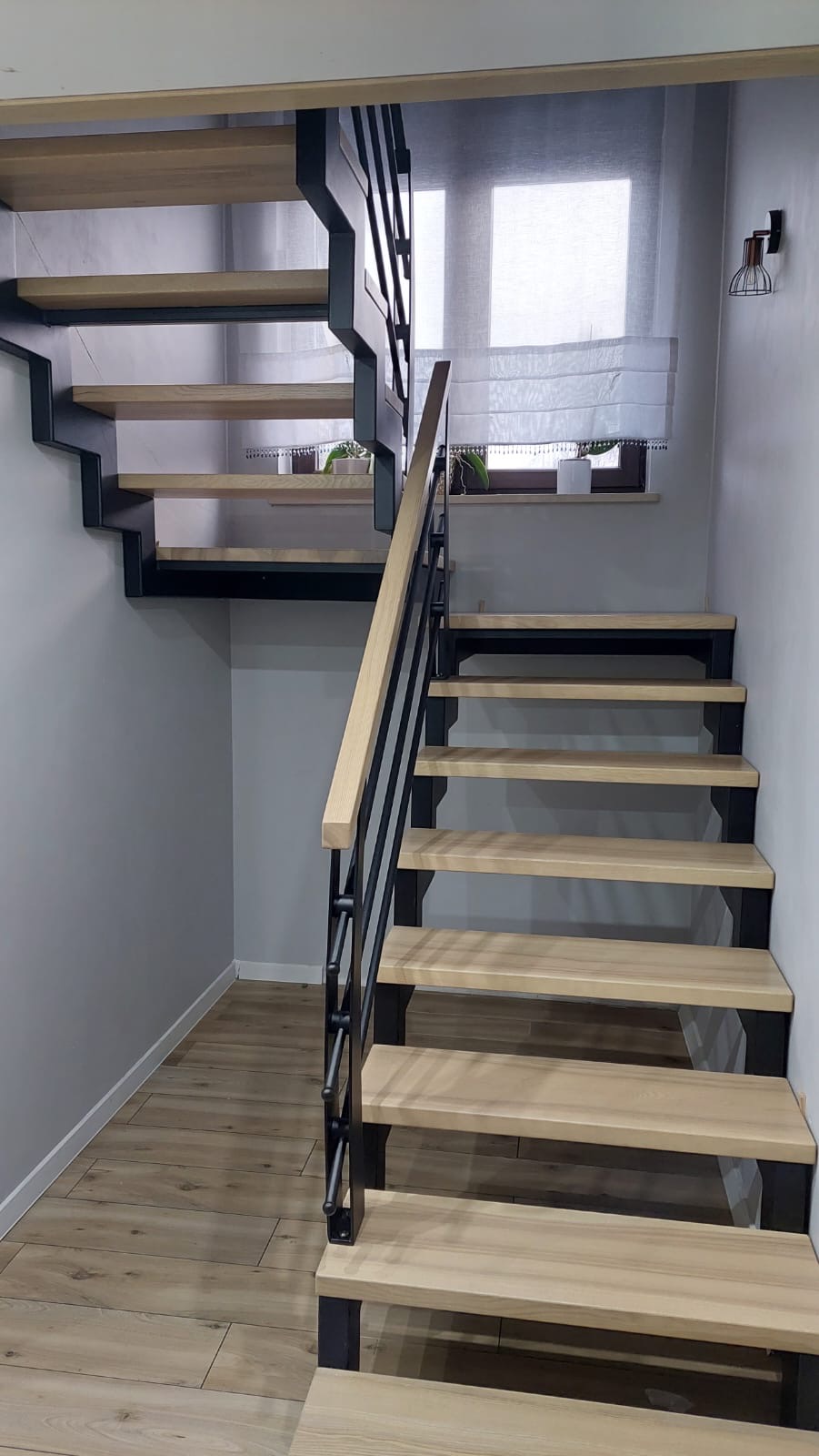 Nowoczesne schody drewniane z czarną metalową konstrukcją i drewnianą balustradą, oświetlone światłem dziennym wpadającym przez okno z roletą rzymską.