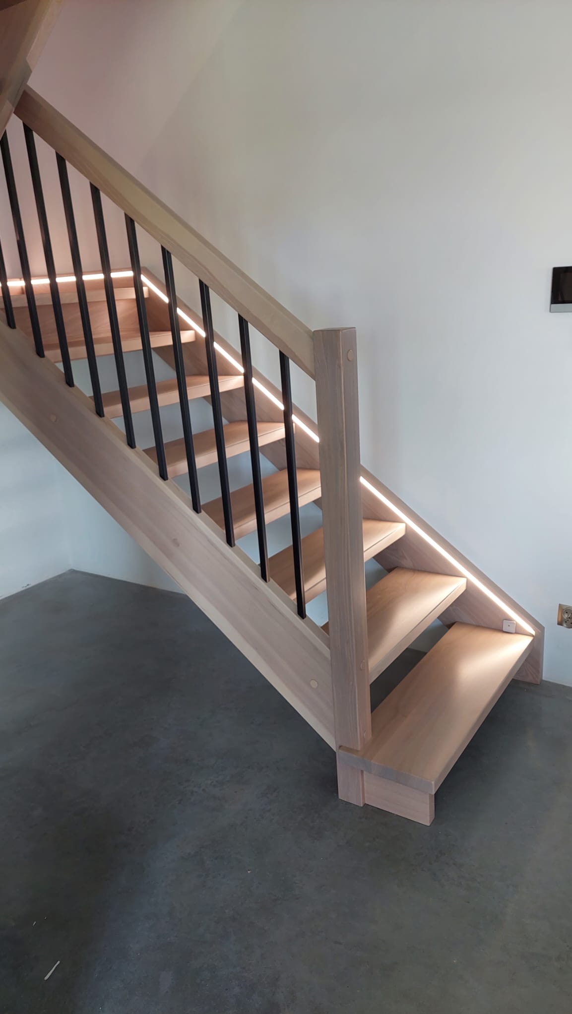 Jasne, drewniane schody z czarnymi balustradami i subtelnym oświetleniem LED wzdłuż stopni i poręczy.