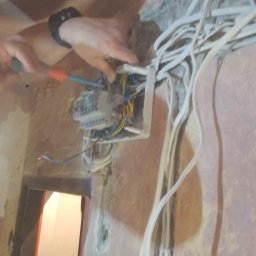 Sławomir Wajman - Cztery Kąty - Elektryk z zegarkiem na ręku montuje skrzynkę elektryczną z licznymi przewodami w ścianie podczas remontu mieszkania, widoczny fragment futryny drzwi.