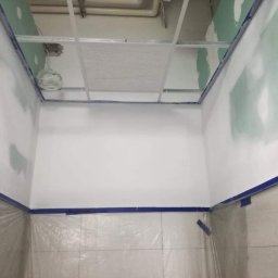 Sławomir Wajman - Cztery Kąty - Wnętrze w trakcie prac wykończeniowych: ściany po wstępnym szpachlowaniu, zabezpieczone niebieską taśmą malarską i folią ochronną na podłodze, widoczna konstrukcja sufitu podwieszanego z przewodami.