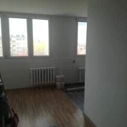 Sławomir Wajman - Cztery Kąty - Pokój po odświeżeniu: świeżo pomalowane białe ściany, drewniana podłoga, grzejnik pod oknem, widoczne wiadro z farbą i akcesoria malarskie, fragment drabiny malarskiej.