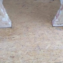 Sławomir Wajman - Cztery Kąty - Surowa płyta OSB ułożona na podłodze pomiędzy dwoma ościeżnicami w trakcie prac remontowych, widoczne ślady zaprawy na ościeżnicach.