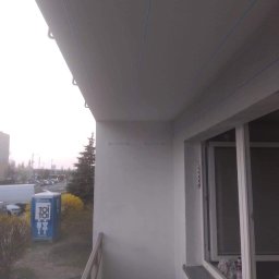 Sławomir Wajman - Cztery Kąty - Biały sufit balkonu z widocznymi niebieskimi liniami i uchwytami, widok na zewnątrz z toaletą przenośną Toi Toi i drzewami w tle.