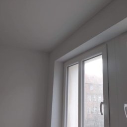 Sławomir Wajman - Cztery Kąty - Świeżo pomalowane, białe wnętrze pokoju z widokiem na budynek przez okno z białymi ramami.