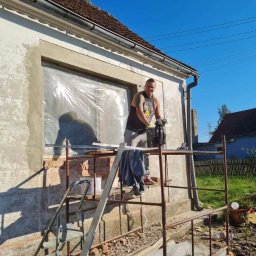 Sławomir Wajman - Cztery Kąty - Pracownik na rusztowaniu przygotowuje ścianę budynku w Skarszynie do nałożenia nowej warstwy, okno zabezpieczone folią, widoczne narzędzia i materiały budowlane.