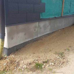 Sławomir Wajman - Cztery Kąty - Fragment elewacji budynku z częściowo ułożonymi czarnymi płytami elewacyjnymi, widoczna folia wiatroizolacyjna i podmurówka z izolacją przeciwwilgociową, teren wokół budynku w stanie surowym.