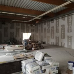 Sławomir Wajman - Cztery Kąty - Wnętrze w trakcie prac wykończeniowych; ściany przygotowane do szpachlowania z widocznymi fragmentami płyt karton-gips, worki z zaprawą na paletach, widoczna osoba w oddali.