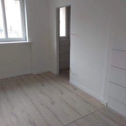 Sławomir Wajman - Cztery Kąty - Wnętrze po remoncie: świeżo pomalowane białe ściany, jasne panele podłogowe i widok na wykafelkowaną łazienkę przez otwarte drzwi.