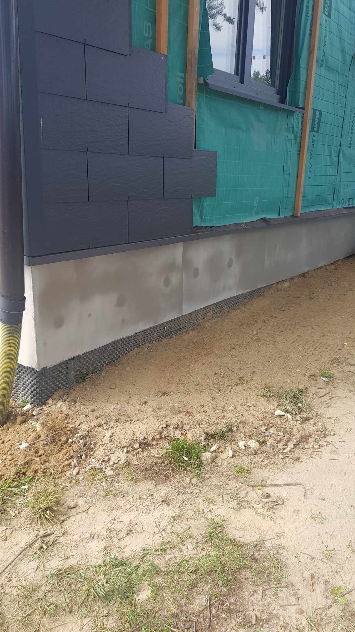 Fragment elewacji budynku z częściowo ułożonymi czarnymi płytami elewacyjnymi, widoczna folia wiatroizolacyjna i podmurówka z izolacją przeciwwilgociową, teren wokół budynku w stanie surowym.