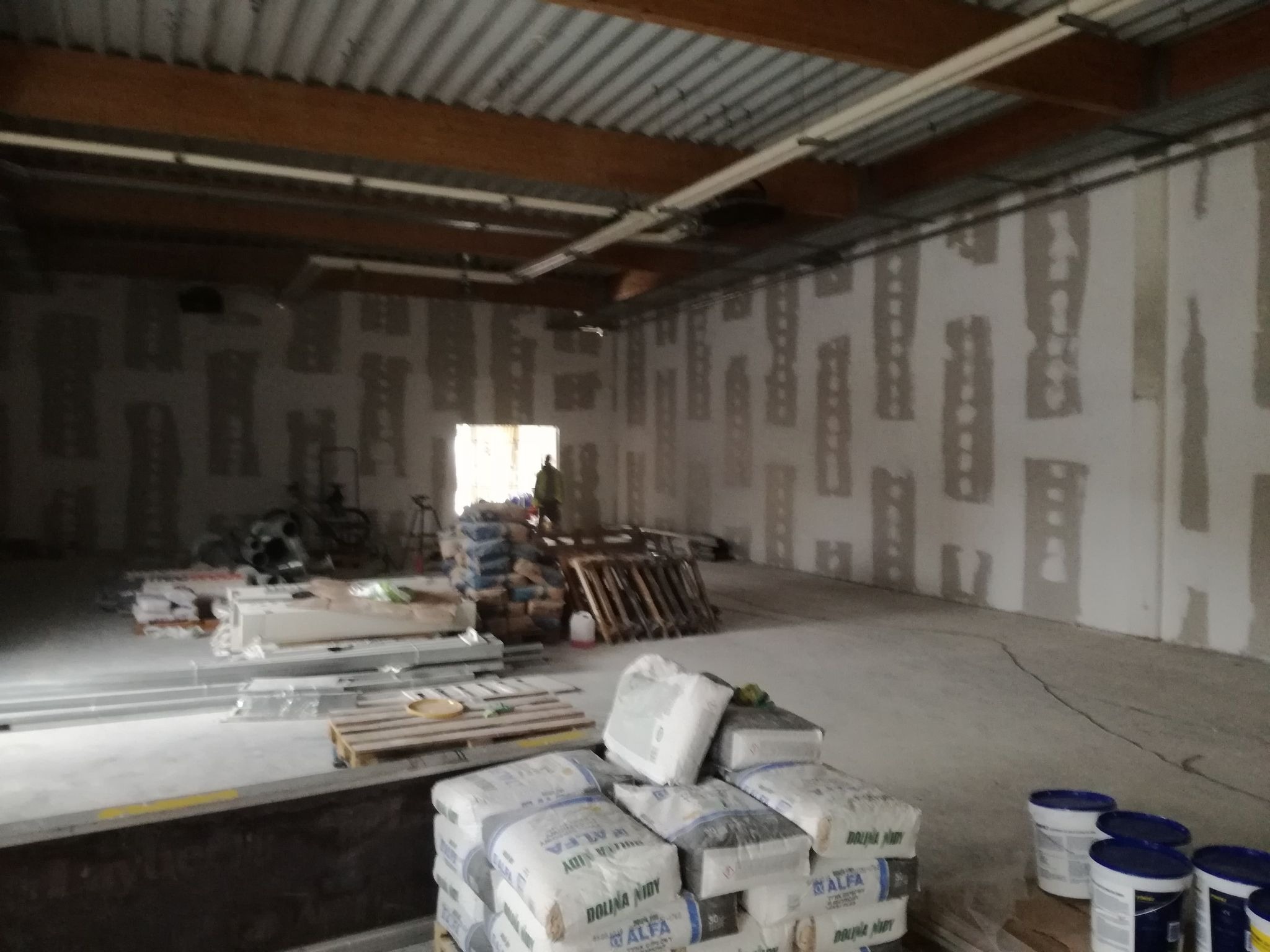 Wnętrze w trakcie prac wykończeniowych; ściany przygotowane do szpachlowania z widocznymi fragmentami płyt karton-gips, worki z zaprawą na paletach, widoczna osoba w oddali.