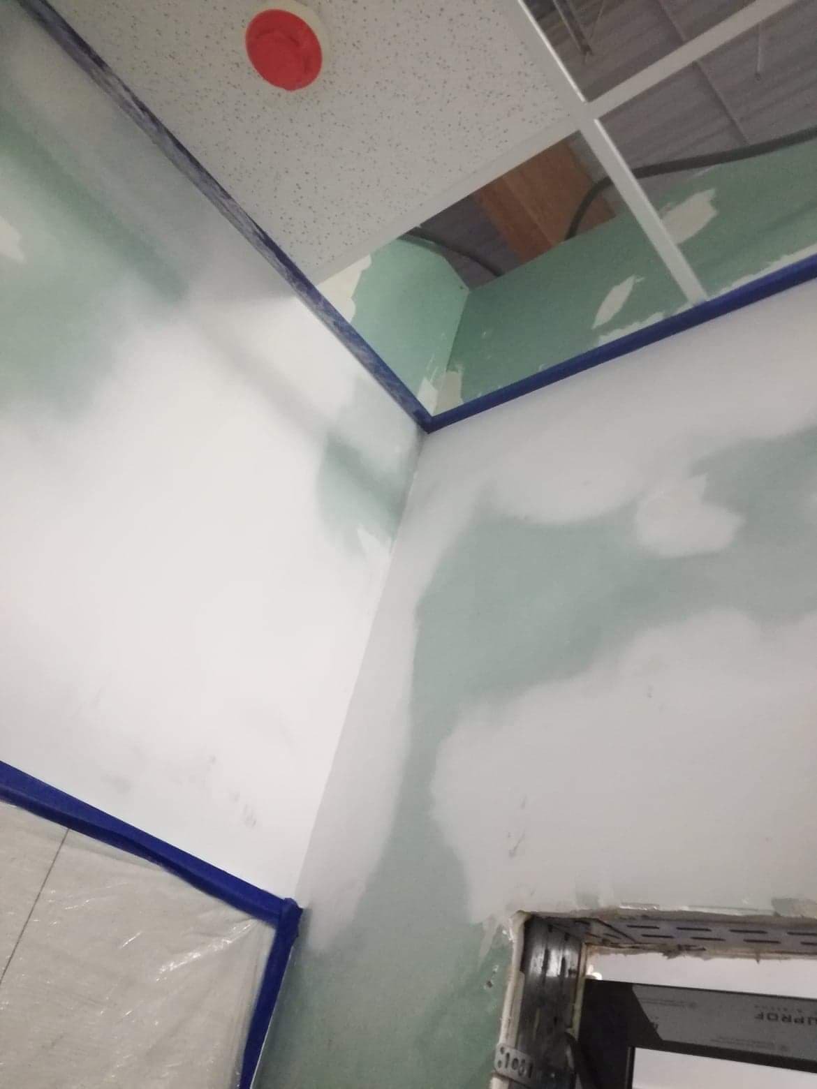 Przygotowanie ścian do malowania: ściany z nałożoną warstwą szpachli, zabezpieczone taśmą malarską przy suficie i folią ochronną przy podłodze, widoczne ślady po szpachlowaniu.