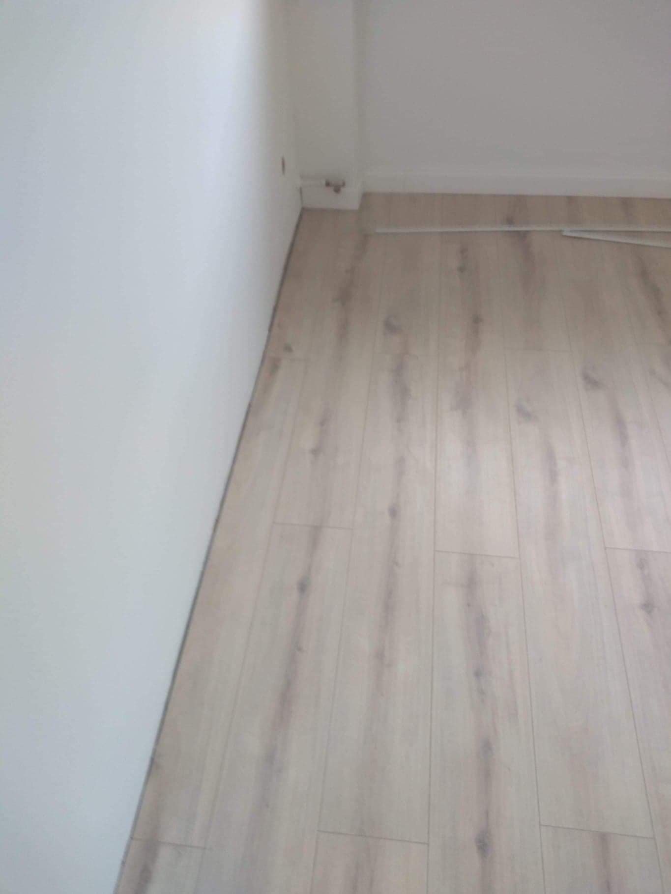 Jasne panele podłogowe ułożone wzdłuż białej ściany i listwy przypodłogowej, widoczne delikatne łączenia między panelami.