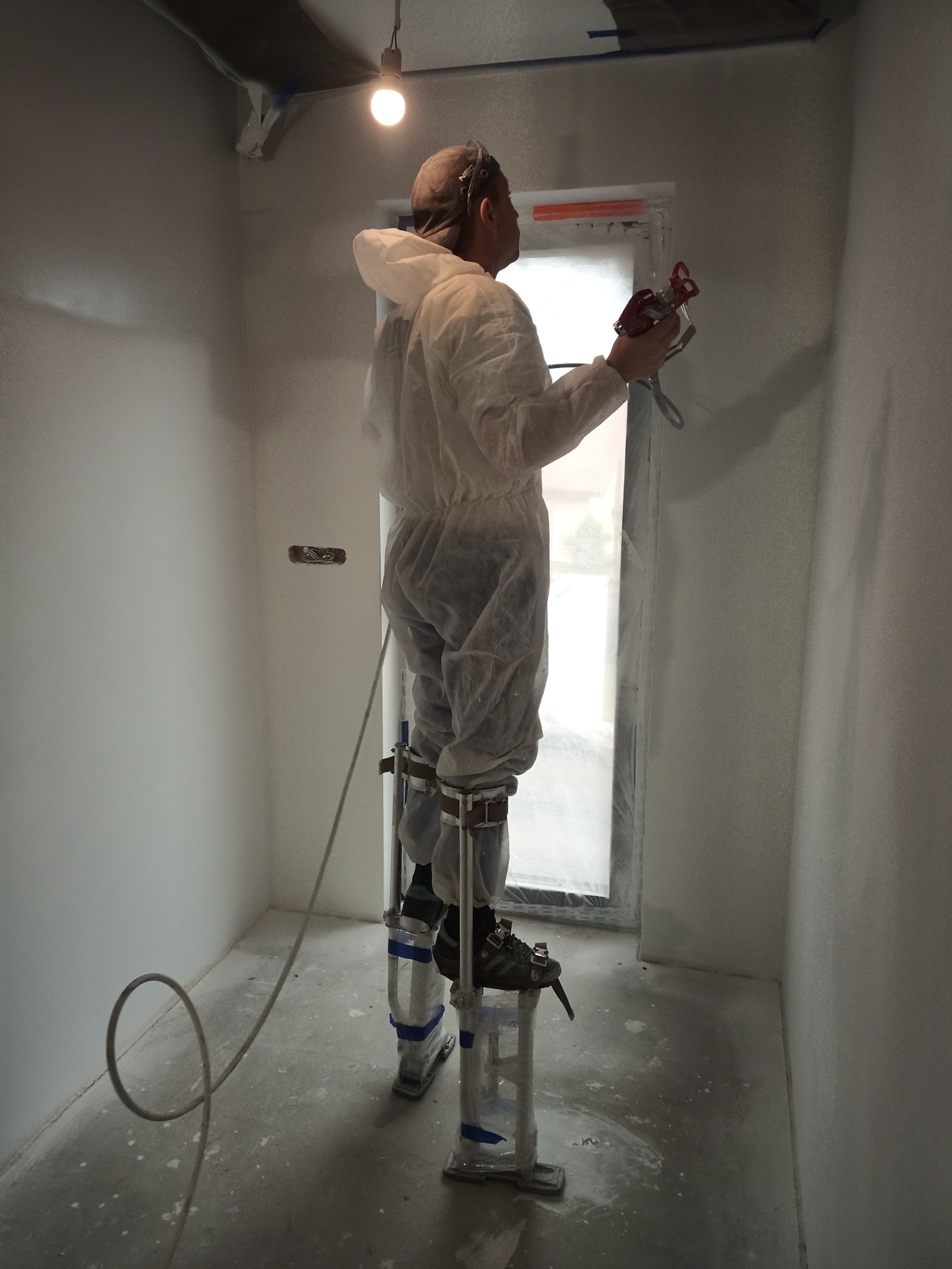 Pracownik w kombinezonie ochronnym i na szczudłach, maluje ściany w pomieszczeniu pistoletem natryskowym, widoczny kabel i osprzęt, okno zabezpieczone folią.