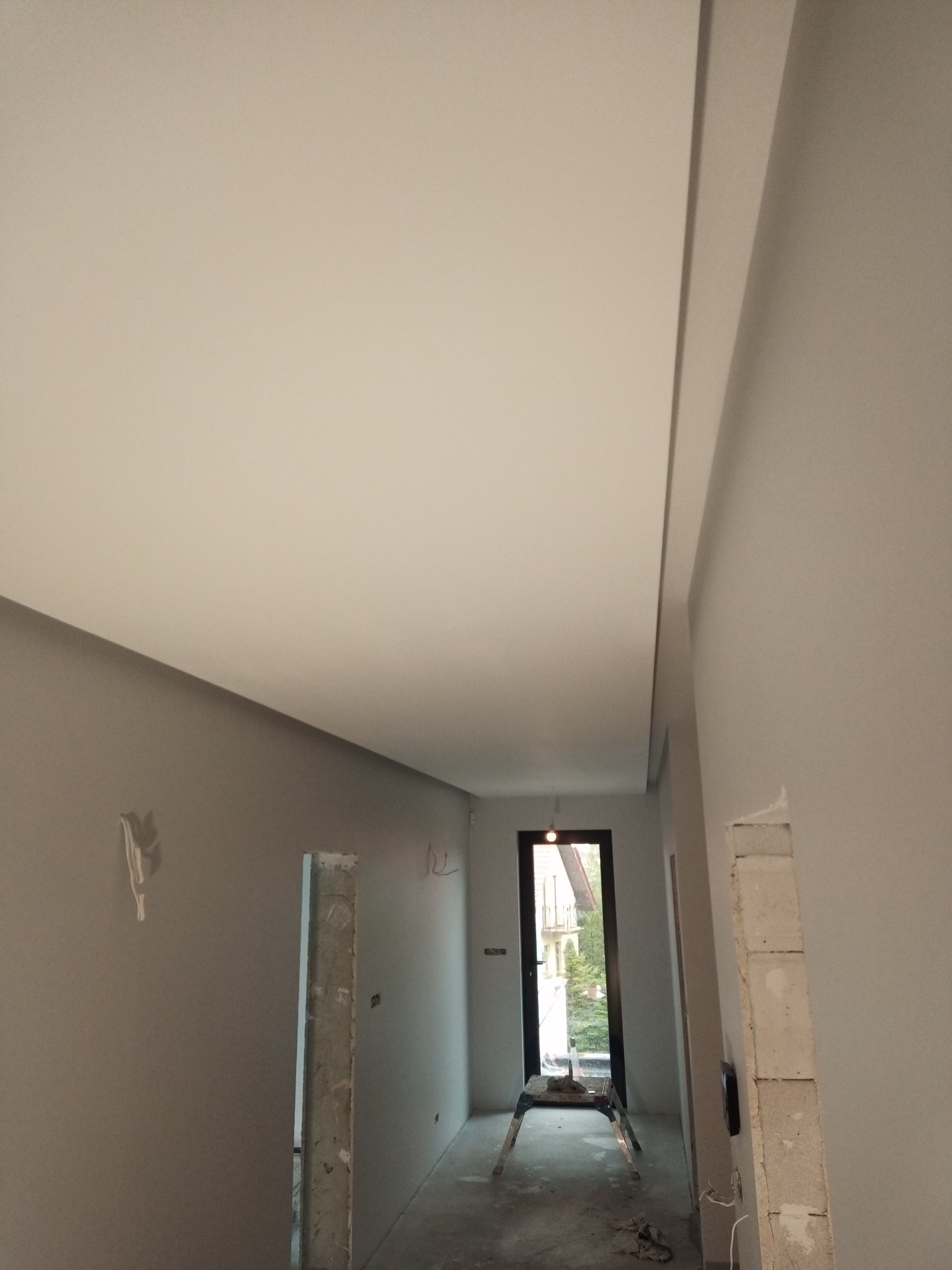 Wnętrze w trakcie wykańczania: pomalowane ściany w odcieniach szarości, sufit z płyty karton gips, widoczna drabina malarska przy drzwiach balkonowych i fragmenty niezabezpieczonych otworów...