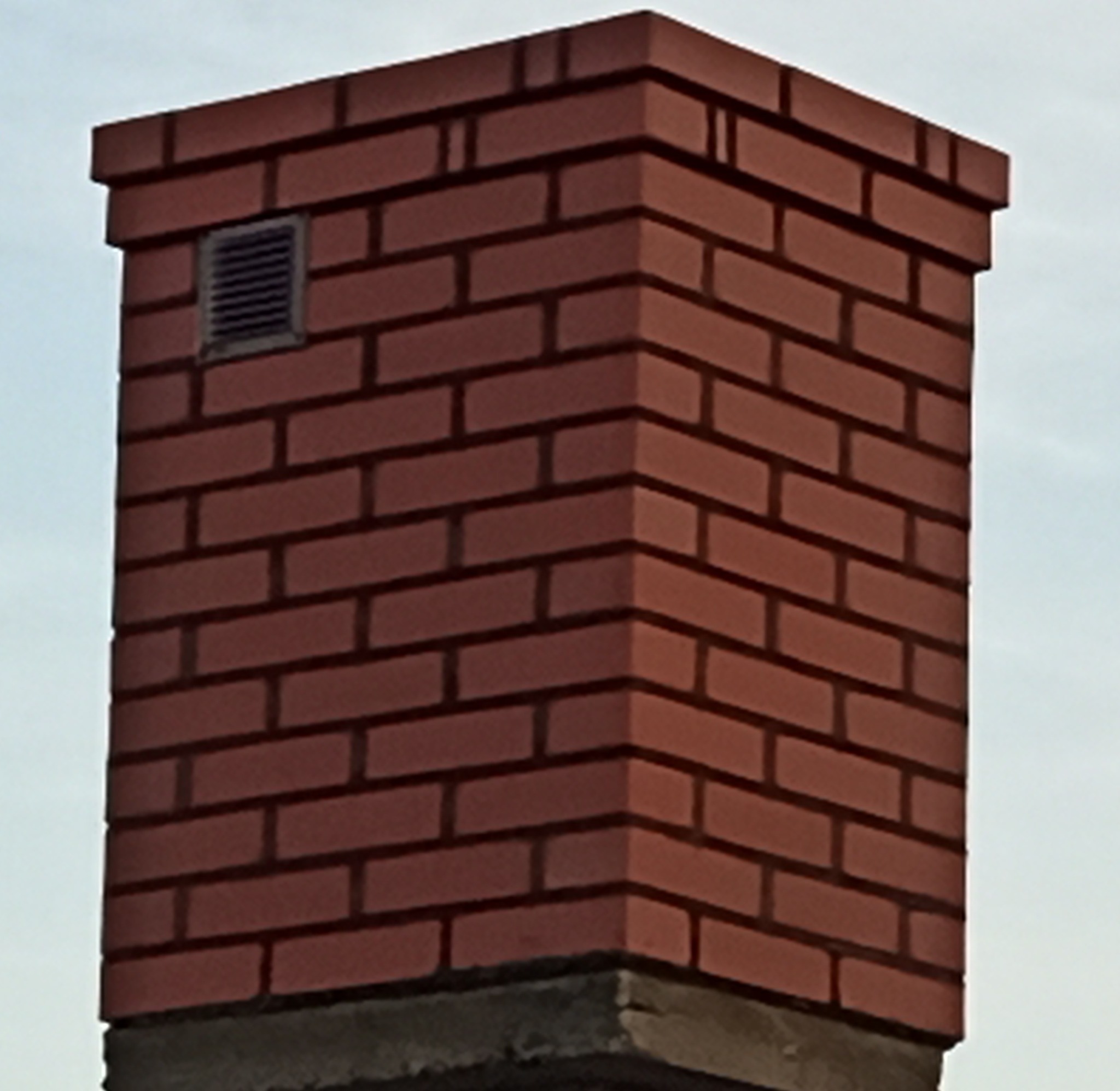 Ceglany komin z kratką wentylacyjną, widoczny od dołu na tle jasnego nieba.