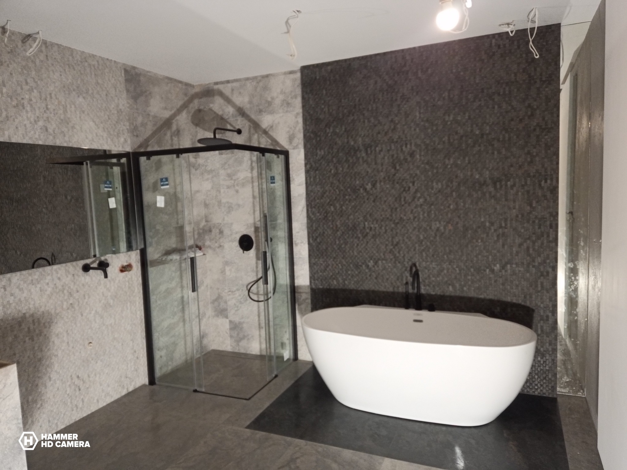 Nowoczesna łazienka z kabiną prysznicową typu walk-in i wolnostojącą wanną na tle mozaiki ściennej w odcieniach szarości, widoczne niedokończone prace elektryczne przy suficie.