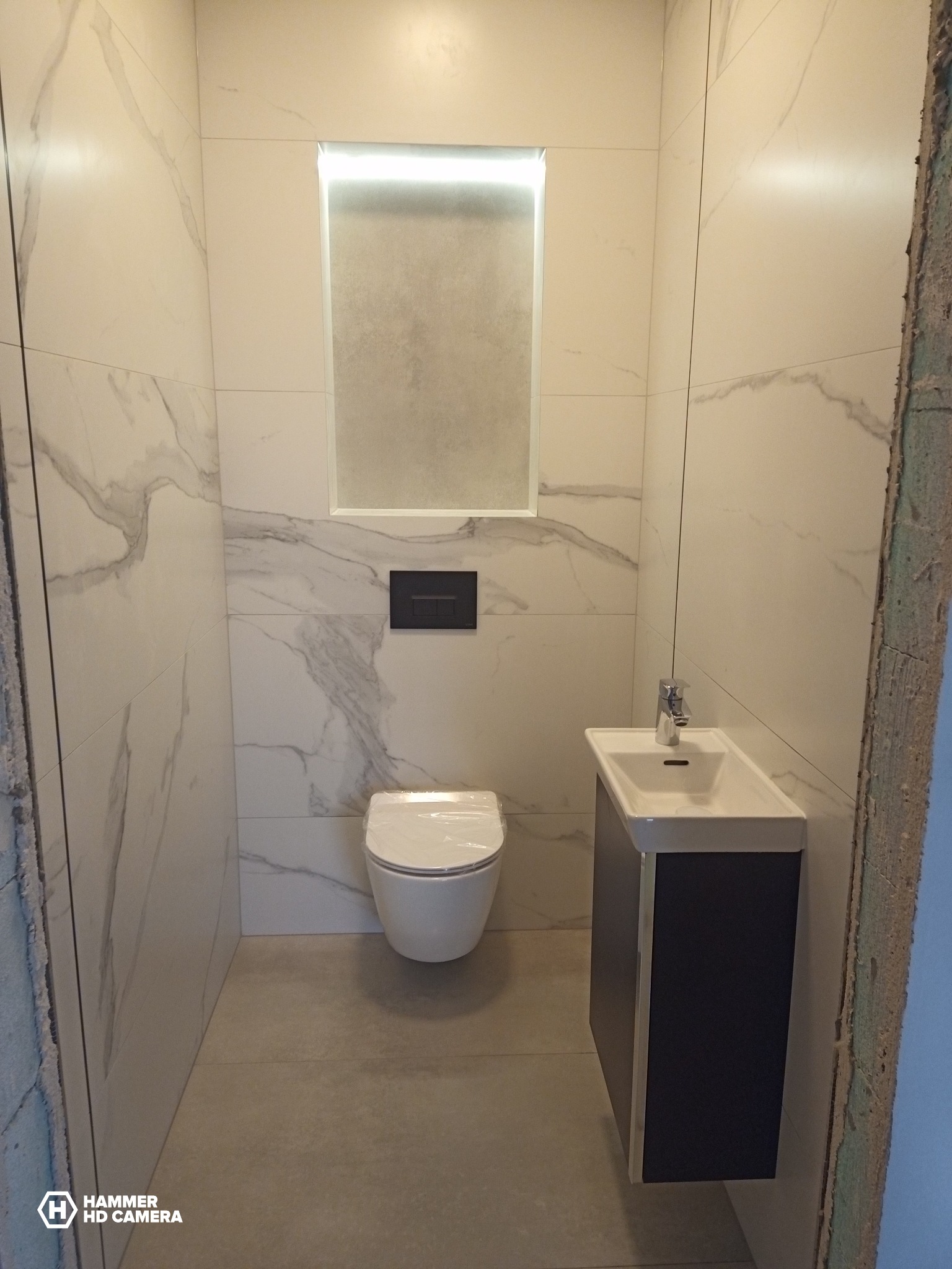 Nowoczesna łazienka w trakcie wykańczania, widoczne marmurowe płytki, podwieszana toaleta, umywalka z szafką i podświetlane lustro; surowy fragment ściany po prawej stronie sugeruje trwający remont.