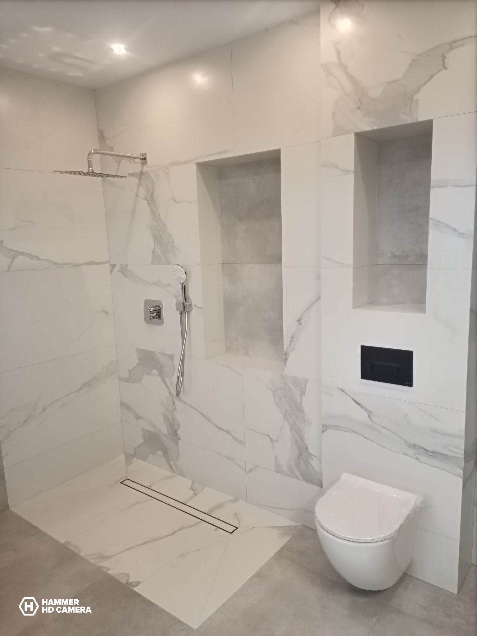 Nowoczesna łazienka wykończona białymi płytkami imitującymi marmur, z kabiną prysznicową typu walk-in, deszczownicą, wnękami w ścianie i podwieszaną toaletą z czarnym przyciskiem.