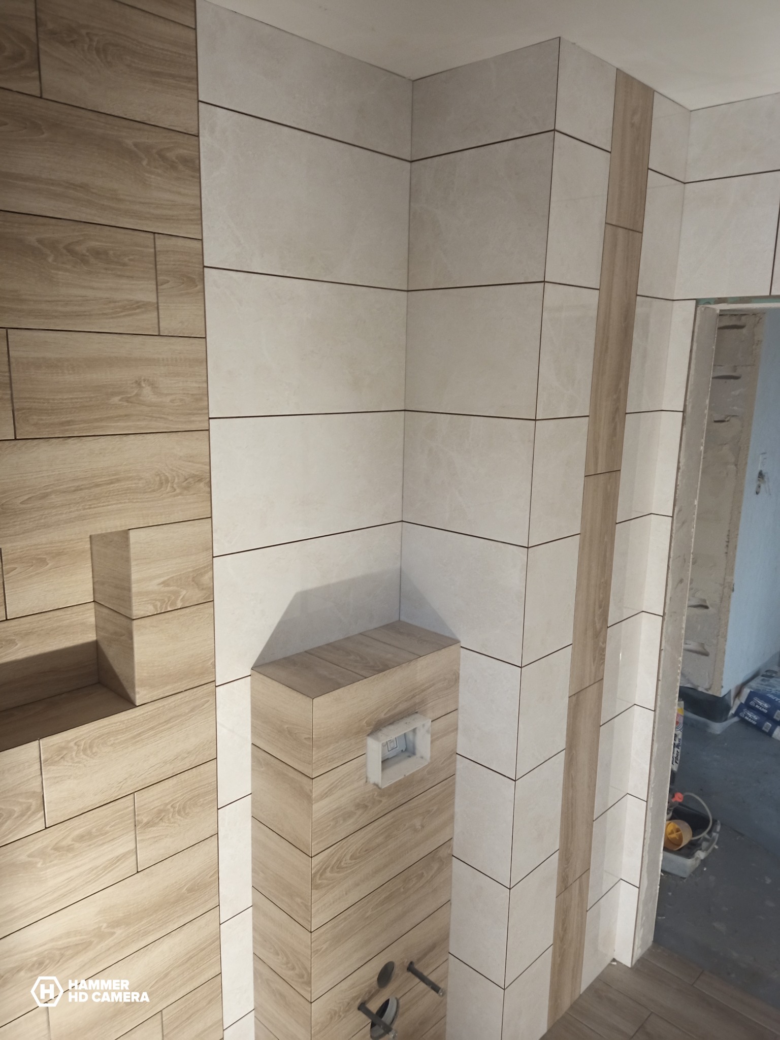 Wykończona łazienka z kombinacją płytek imitujących drewno i jasnych płytek ceramicznych, zabudowa stelaża WC w drewnie, widoczny fragment wejścia do łazienki w trakcie prac wykończeniowych.