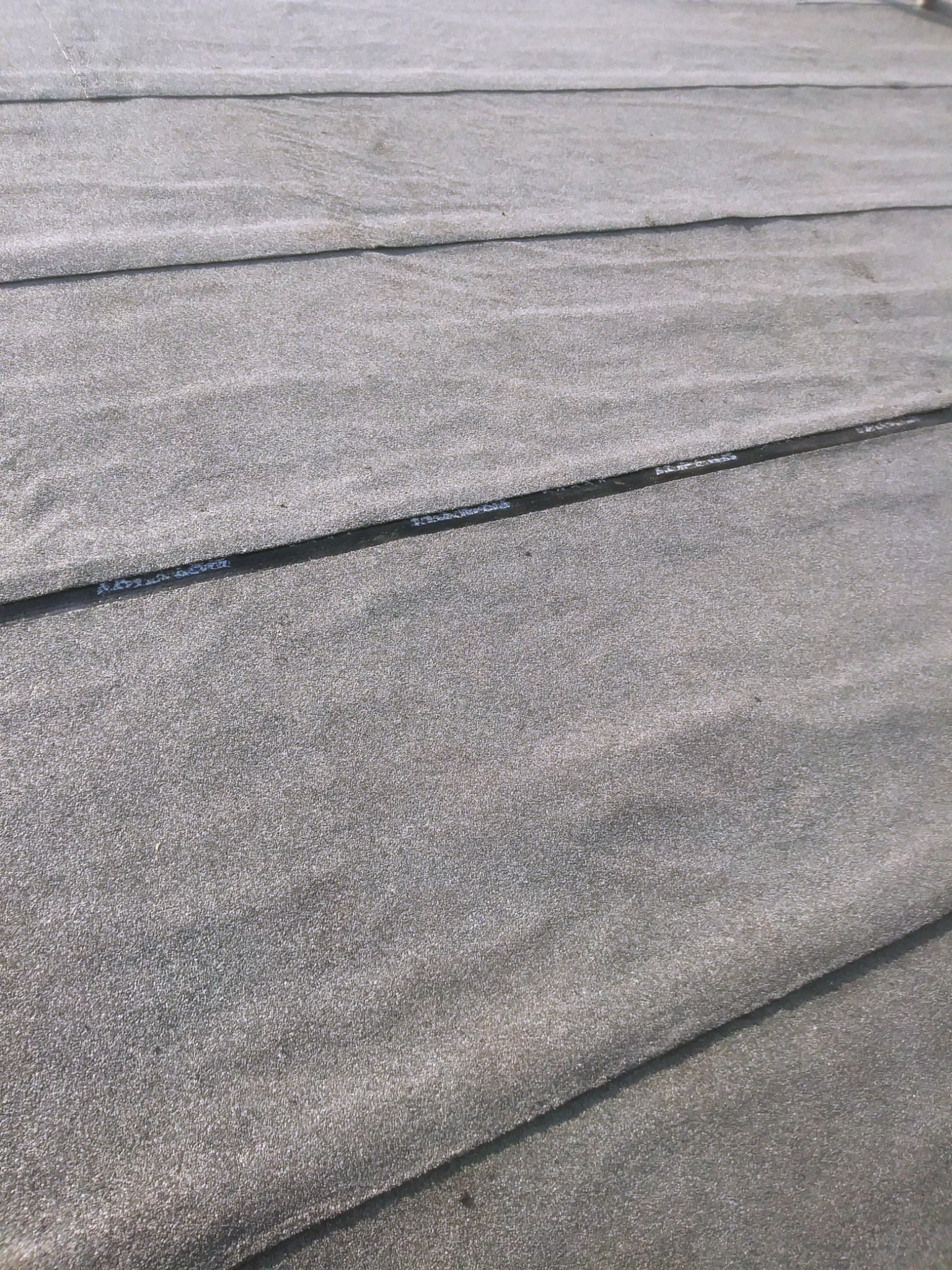 Szary, płaski dach pokryty papą z widocznymi łączeniami i napisami na spodniej warstwie.