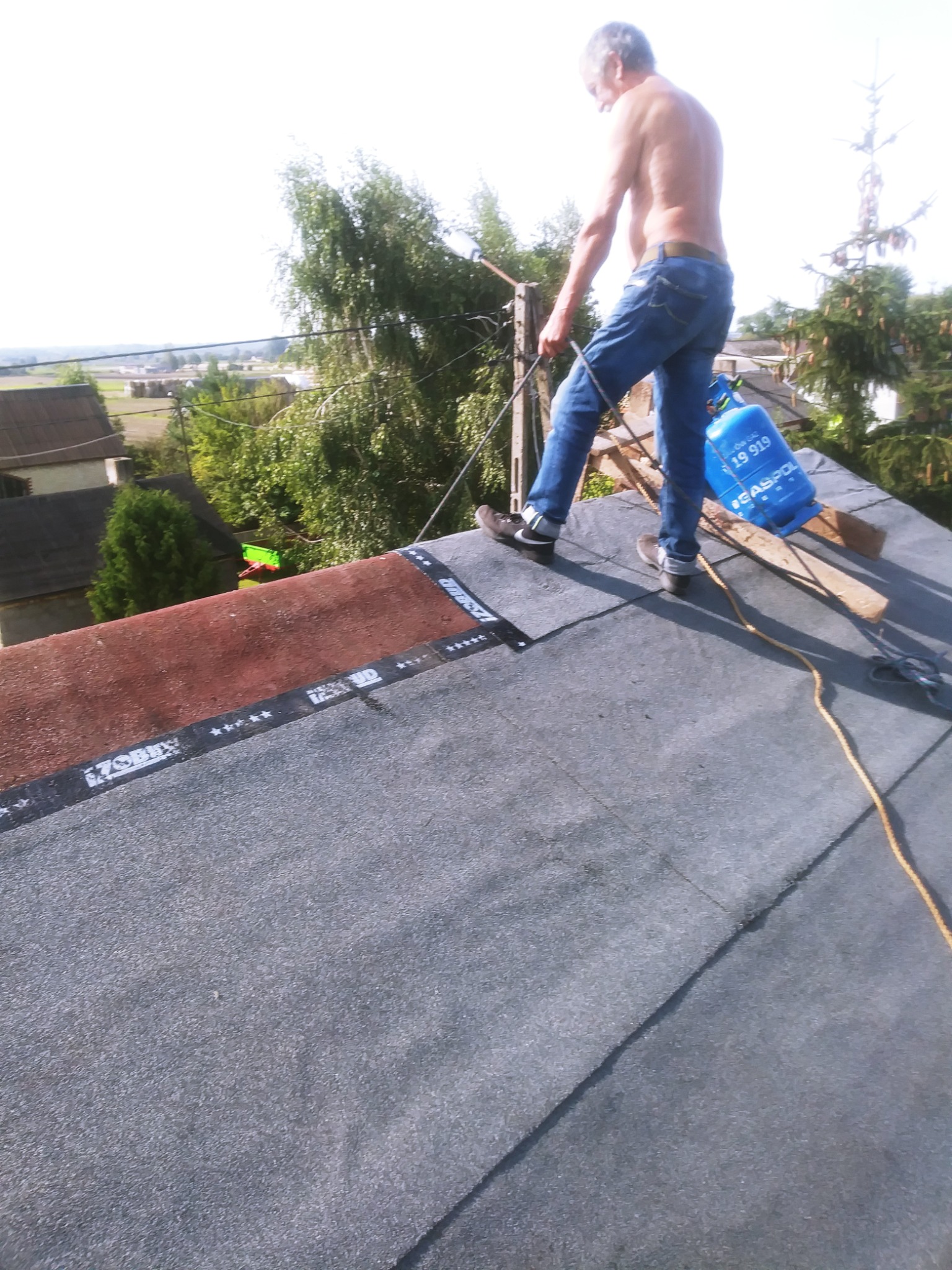 Dekarz bez koszulki z butlą gazową na dachu pokrytym papą termozgrzewalną, zabezpieczony liną, w tle zabudowa jednorodzinna i drzewa.