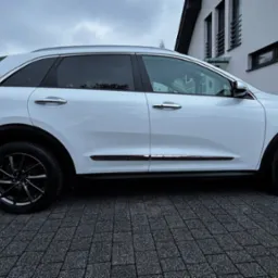 Biały samochód typu SUV, Kia Niro, zaparkowany na kostce brukowej przed domem, widok z boku, czarne felgi, lekko pochmurne niebo.
