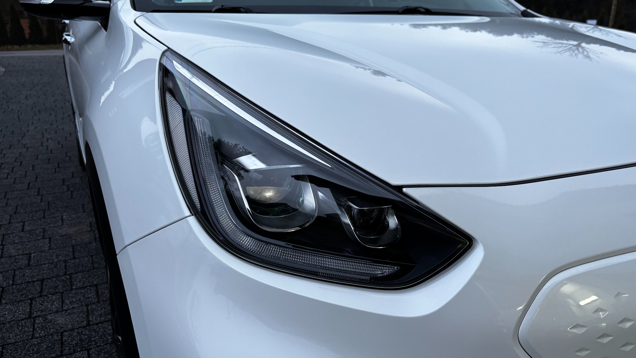 Biały samochód z bliska, widoczny reflektor z detalami i odblaskami światła, karoseria po umyciu lśni czystością.