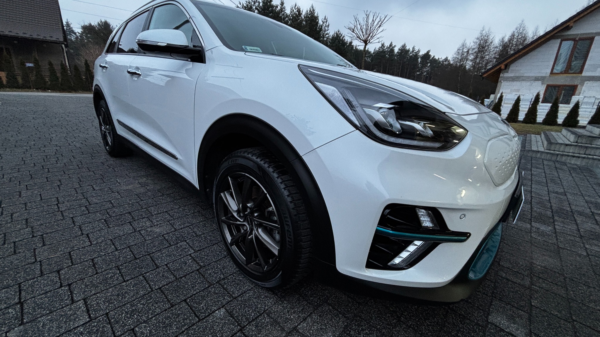 Biały samochód elektryczny Kia Niro zaparkowany na kostce brukowej, widok z niskiej perspektywy na przednie koło i reflektor, w tle dom w trakcie budowy i drzewa.