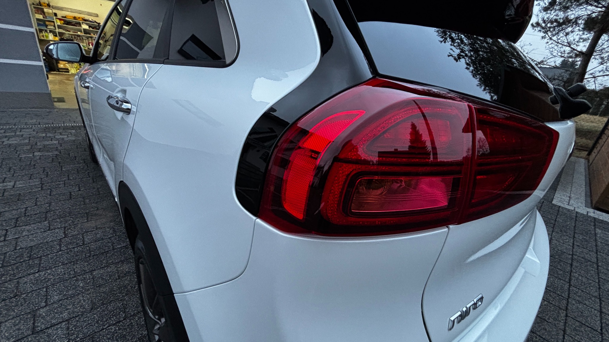 Błyszczący, biały samochód Kia Niro po umyciu, widoczny z tyłu, z wyraźnie odbijającym światło czerwonym kloszem lampy.