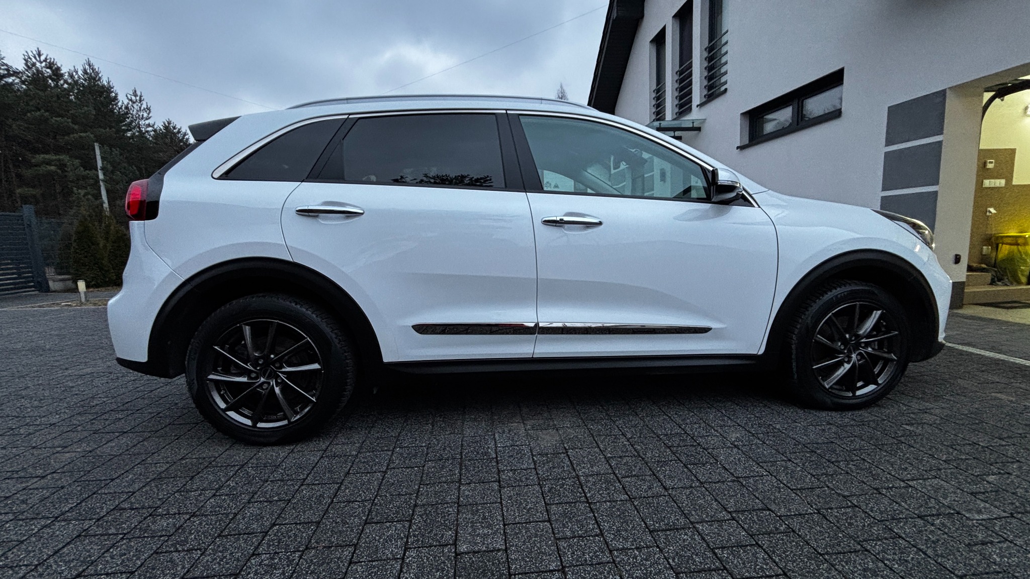 Biały samochód typu SUV, Kia Niro, zaparkowany na kostce brukowej przed domem, widok z boku, czarne felgi, lekko pochmurne niebo.
