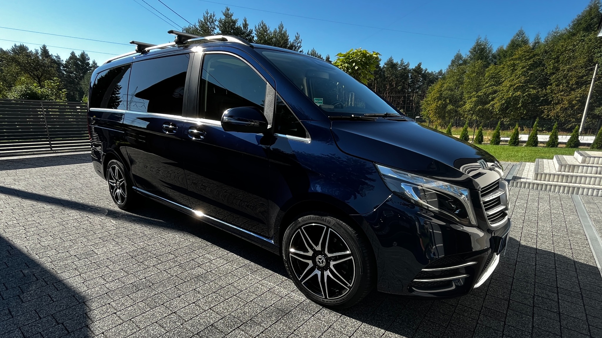 Błyszczący, granatowy van Mercedes-Benz V-klasy z relingami dachowymi, zaparkowany na kostce brukowej w słoneczny dzień.