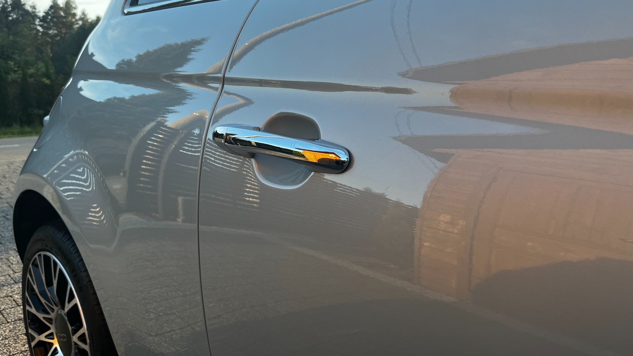 Błyszczący lakier i chromowana klamka na boku szarego samochodu, z widocznymi refleksami otoczenia.