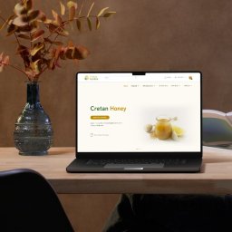 Edamame - Laptop wyświetla stronę internetową o miodzie kreteńskim. Obok wazon z gałązkami i kobieta czytająca książkę. Stonowana kolorystyka, ciepłe światło.