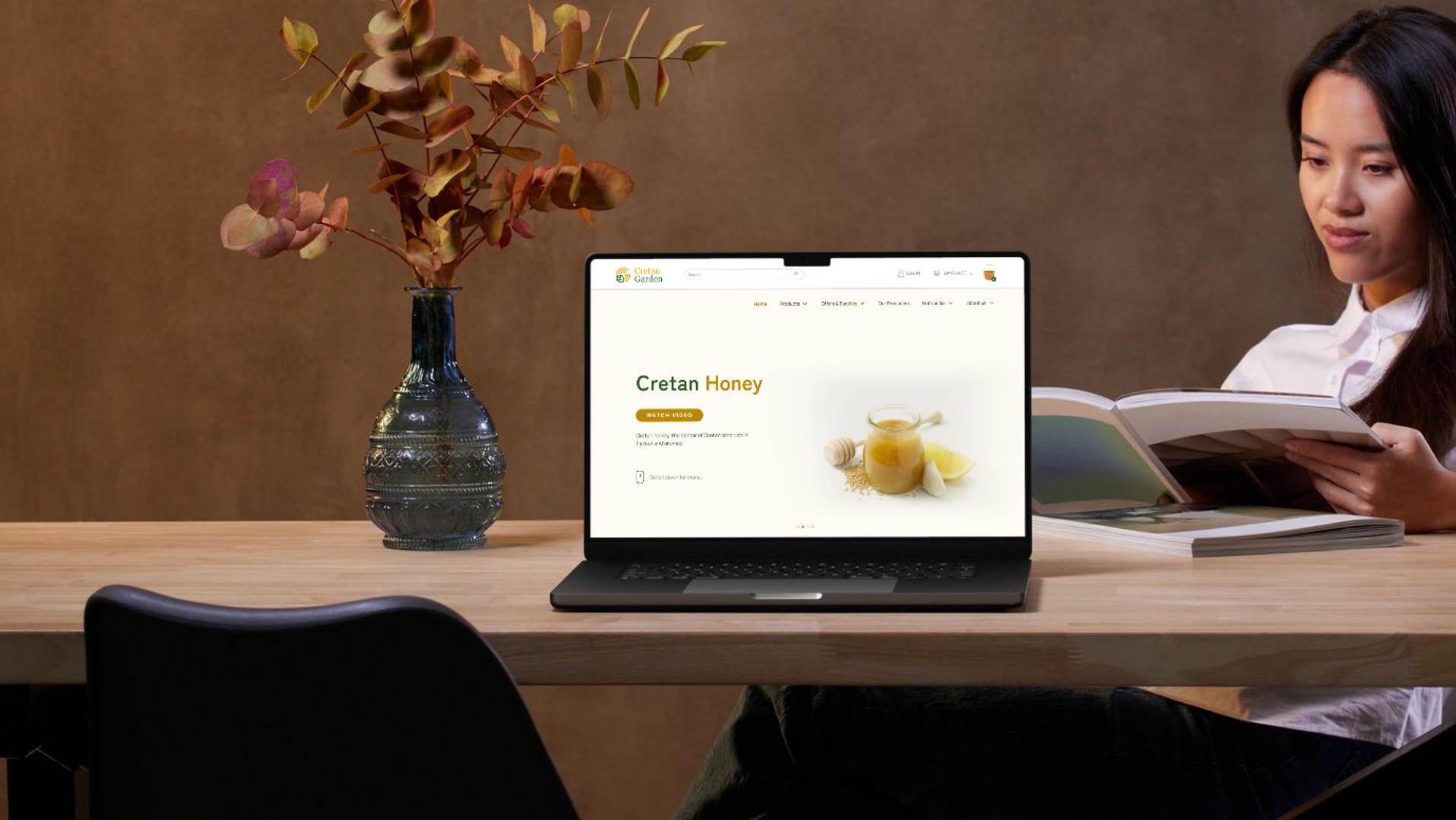 Laptop wyświetla stronę internetową o miodzie kreteńskim. Obok wazon z gałązkami i kobieta czytająca książkę. Stonowana kolorystyka, ciepłe światło.