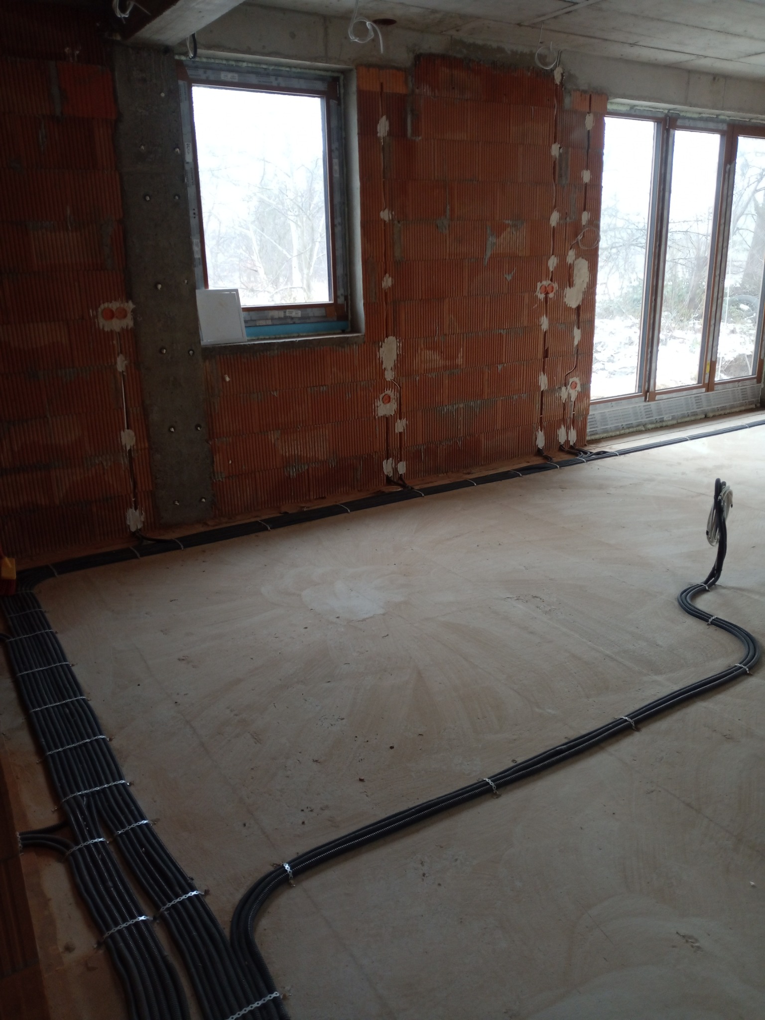 Wnętrze w stanie surowym z widocznymi przewodami elektrycznymi ułożonymi na podłodze i poprowadzonymi w bruzdach w ścianach z cegły, przygotowanymi pod tynk.