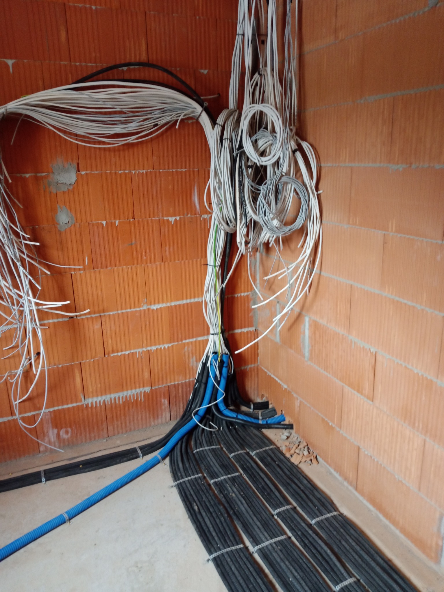Surowe ściany z cegły dziurawki z widoczną wiązką przewodów elektrycznych, częściowo ułożonych w peszlach, w trakcie montażu.