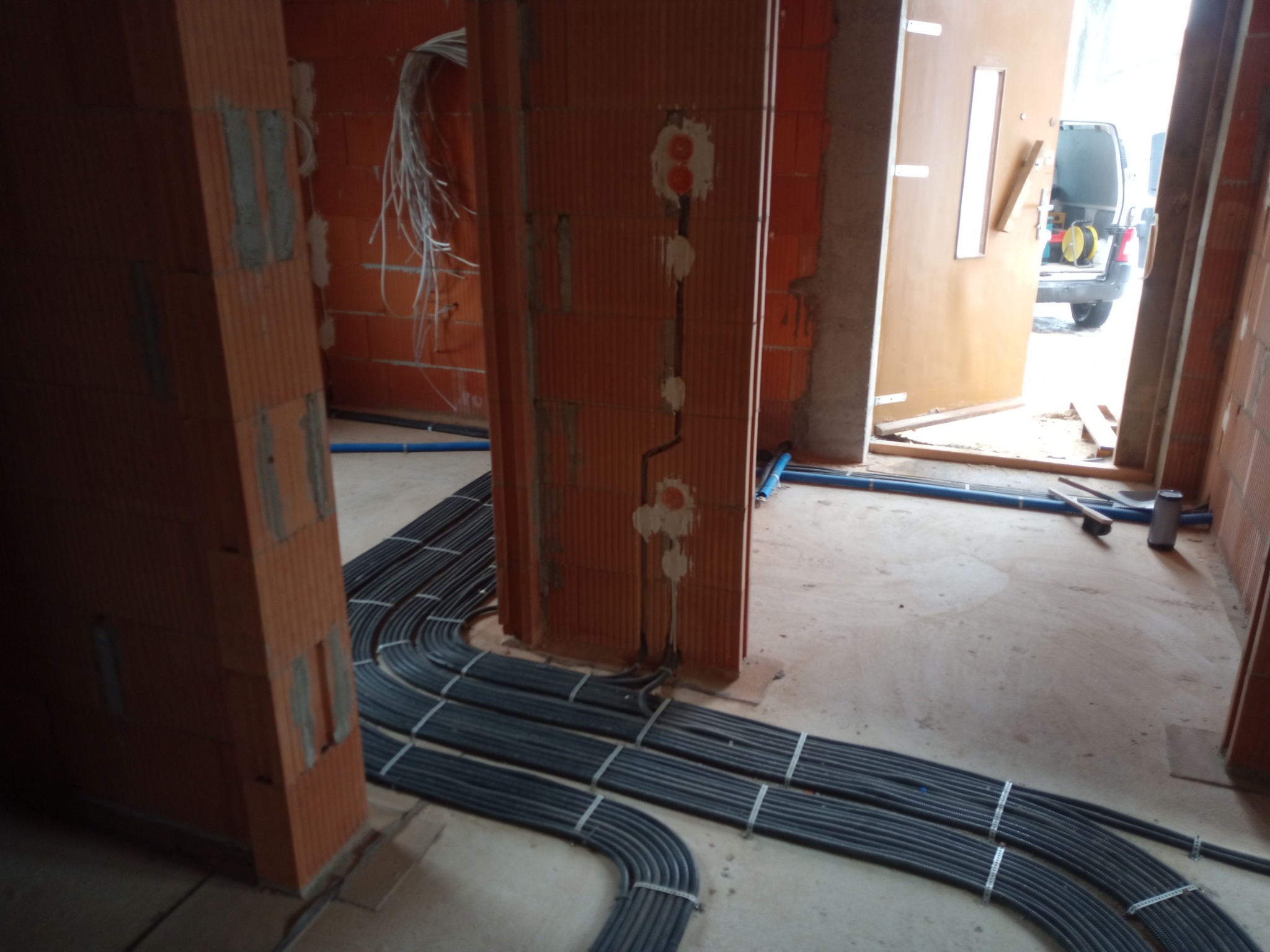 Wnętrze w stanie surowym z widocznymi przewodami elektrycznymi ułożonymi w rurach na podłodze, pionowe bruzdy w ścianie z wyprowadzonymi kablami, w tle otwarte drzwi i zaparkowany samochód dostawczy.