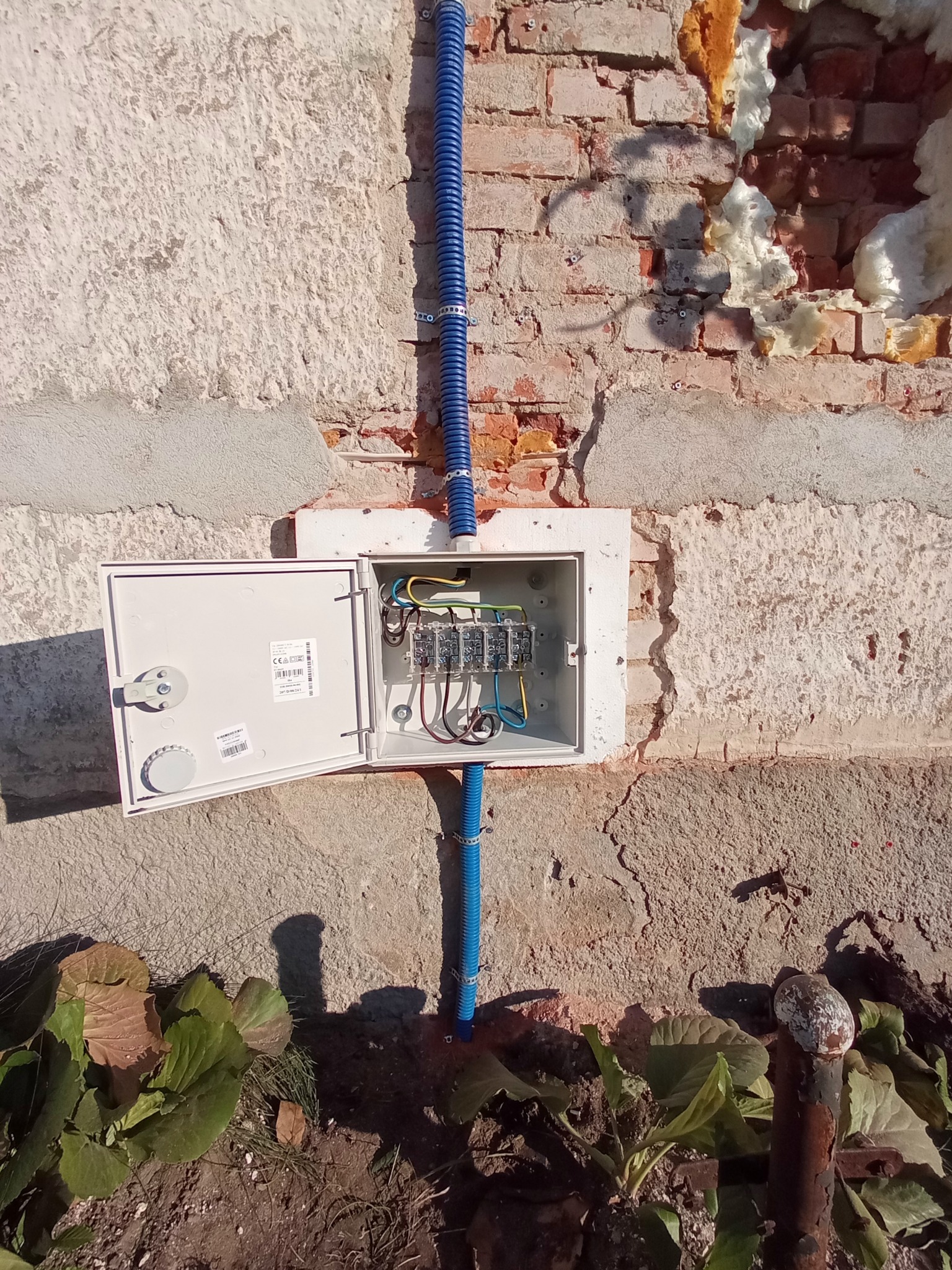 Otwarta skrzynka elektryczna zamontowana na nierównej ścianie z cegły i tynku, z widocznymi przewodami i niebieskimi rurami karbowanymi.