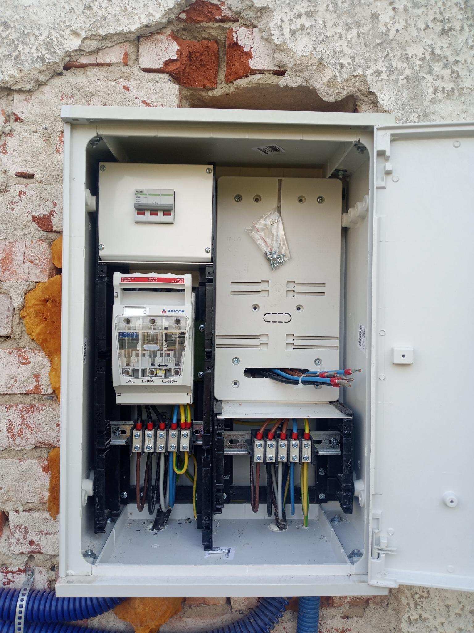 Otwarta skrzynka elektryczna z zainstalowanym licznikiem i zabezpieczeniami na tle ściany z widoczną cegłą i pianką montażową.