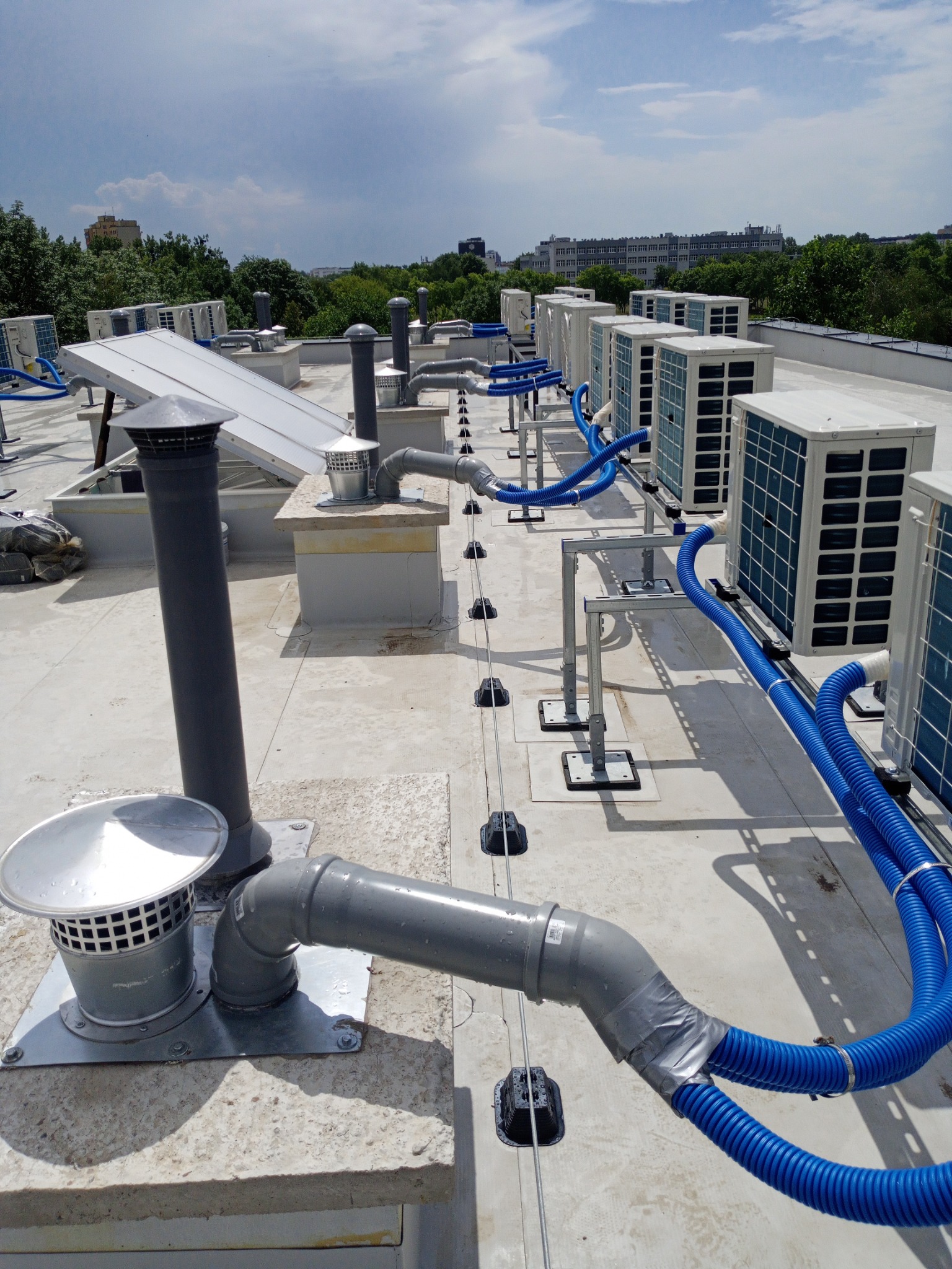 Instalacja wentylacyjna i klimatyzacyjna na płaskim dachu budynku, widoczne rury wentylacyjne, kominki, panele słoneczne oraz szereg jednostek zewnętrznych klimatyzacji, niebieskie przewody...
