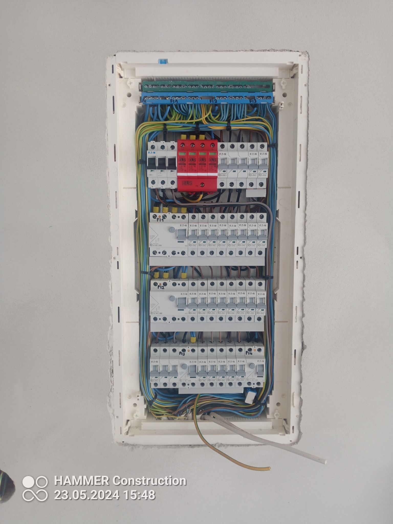 Rozdzielnica elektryczna w białej obudowie, z widocznymi bezpiecznikami, przewodami w kolorach niebieskim, żółtym i zielonym, zamontowana w surowej ścianie.