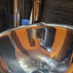 Błyszcząca, metalowa umywalka z widocznym odpływem i odbiciem pomarańczowej ściany oraz fragmentu łazienki, obok bateria z nakładką w kształcie uszu kota.