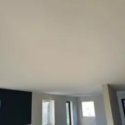 Gładka, jasna powierzchnia sufitu po remoncie w Koszalinie, widoczna z dołu, z oknami w tle. Minimalistyczne wykończenie wnętrza.